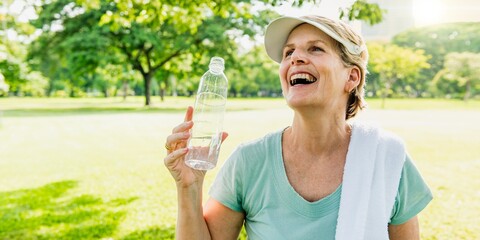 Smiling healthy elderly woman outdoors holding a water bottle, wearing a cap and towel. Enjoying nature and exercise in a sunny park. Smiling elderly woman drinking water in the sunny park.