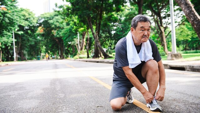 Elderly Asian man tying shoelaces on a park path, preparing for exercise. The Asian man is focused on fitness in the park. Healthy elderly Asian man tying running shoes in the park. - Powered by Adobe