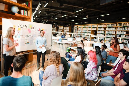 A diverse group in a library setting, with a woman presenting and a man standing by a whiteboard. Students, presentation, diverse group, learning environment. High school students presenting in class.
