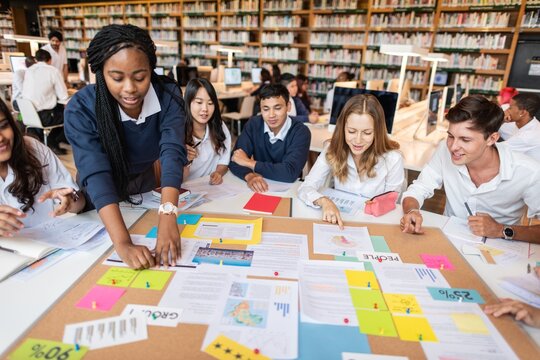 Diverse group of students collaborating in a library, discussing projects, sharing ideas, and working together around a table filled with colorful papers and notes. Young diverse students learning.