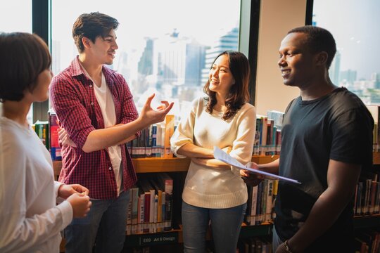 Group of diverse young adults in a library, discussing and smiling. Casual attire, diverse ethnicities, engaging in conversation, standing by bookshelves. Diverse high school students talking. - Powered by Adobe