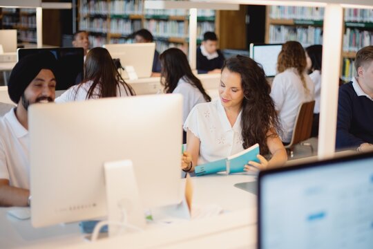 Students studying in a library, with computers and books. Diverse group of students, including an Indian man wearing a turban, focused on their work. High school international students, education.