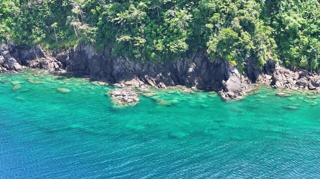 A captivating aerial shot reveals where lush, tree-lined tropical cliffs merge with crystal clear ocean waters at Looc Beach in the Philippines, showcasing rugged rocky shorelines and pristine coast.