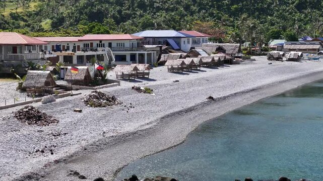 An establishing aerial view over Looc Pebble Beach in Surigao, Philippines, showcasing beach resorts, cozy cottages, and small fishing boats lining the pebble shoreline, perfect for travel visuals.