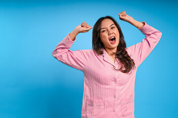 Asian woman in pink pajamas stretching both arms while yawning with mouth wide open, expressing...