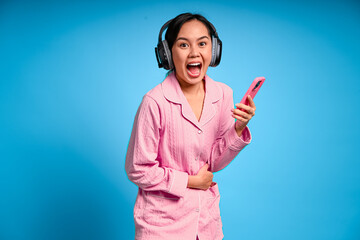 Asian woman in pink pajamas wearing headphones holding her stomach and smartphone while making a...