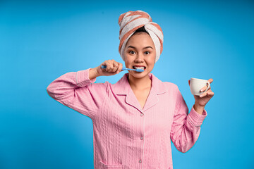 Asian woman in pink pajamas brushing her teeth while holding a white mug, with a towel wrapped on...