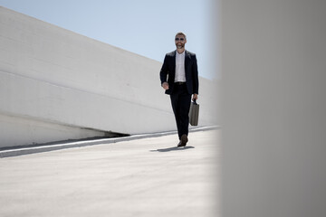Fototapeta premium Business outdoor portrait. Business man walking outdoors. Corporate leader on move. Man in suit with briefcase. Business man going to work. Businessman with briefcase. Business man go to office.