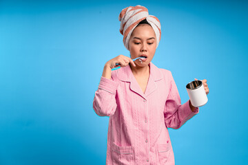 Asian woman in pink pajamas brushing her teeth while holding a white mug, with a towel wrapped on her head, smiling in front of a solid blue background, showing fun morning hygiene routine