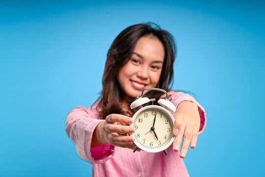 Asian woman in pink pajamas smiling warmly while holding a classic white alarm clock toward the camera, against a solid blue background, symbolizing morning routine and wake-up time