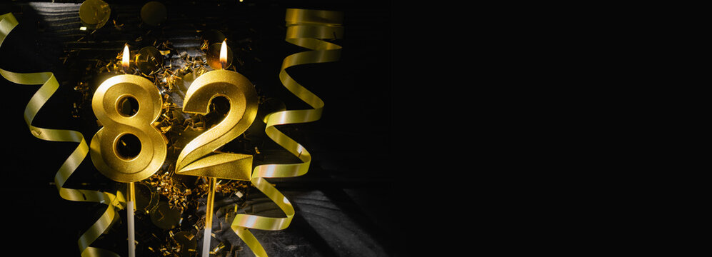 Golden candles number eighty-two burning brightly, celebrating 82nd birthday anniversary. Festive ribbons and confetti on black background, creating elegant celebratory banner.