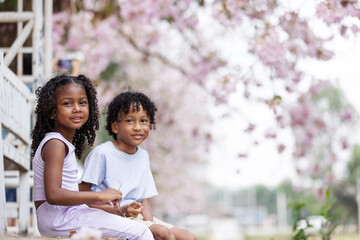 Two children sit on wooden steps, smiling under blooming pink cherry blossoms. The soft, pastel background enhances the warmth of their joyful expressions, creating a serene and cheerful atmosphere.