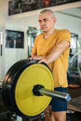 Obraz premium Senior Man with Determined Expression Resting Near Barbell in Gym