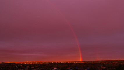 Faint rainbow arching across dramatic sunset sky over rooftops.