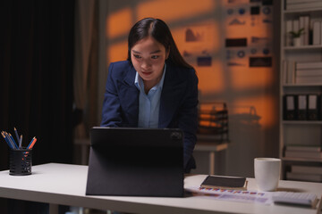 Fototapeta premium Young asian businesswoman concentrating on her work on a tablet in a modern office, working late into the night with warm lighting and business charts visible
