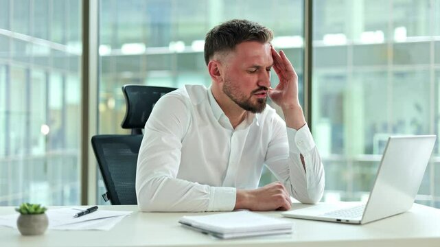 Frustrated businessman experiencing a headache while working on a laptop in a modern office. He rubs his temples, trying to alleviate the pain.