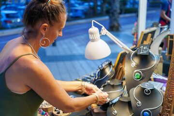 Mature artisan arranging her aluminum wire jewelry display at an outdoor market stall, showcasing...