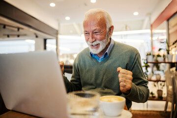 An old man is celebrating victory and looking at laptop in coffee shop.