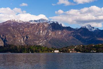 Obraz premium Wide panoramic scene of mountains and Como lake with hillside village nestled at the foot of the hills. Clear blue sky with scattered clouds add to peaceful and scenic atmosphere, Italy