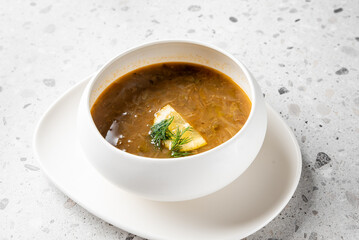 A bowl of rich, hearty soup featuring a creamy texture and a slice of butter on top, garnished with fresh dill, served on a minimalist white plate against a textured background.