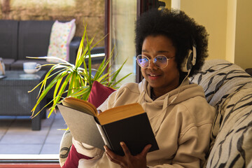 Happy black woman listening music over headphones while reading a book at home