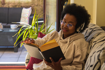 Happy black woman listening music over headphones while reading a book at home