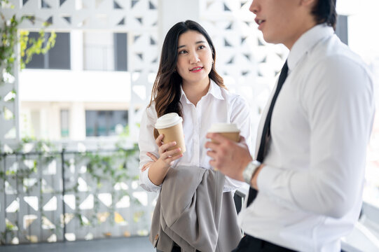 An asian female office worker and male coworker is holding coffee cup while chatting together.