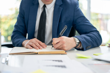 Close up of a hand in blue suit holding pen in left hand and writing or noting down in notebook.