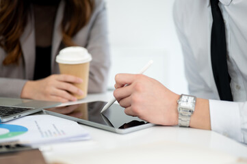 Close up of male hand pointing stylus at tablet showing business data to female coworker in office.