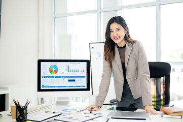Businesswoman or female office worker is leaning over table next to monitor showing data and charts.
