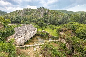 Old stone house in Blagaj village in Bosnia and Herzegovina, Europe. Historic stone building with arched openings over a small river, surrounded by lush greenery and a scenic landscape.