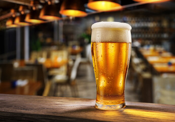 Glass of chilled beer on wooden bar table top and blurred bar interior at the background.
