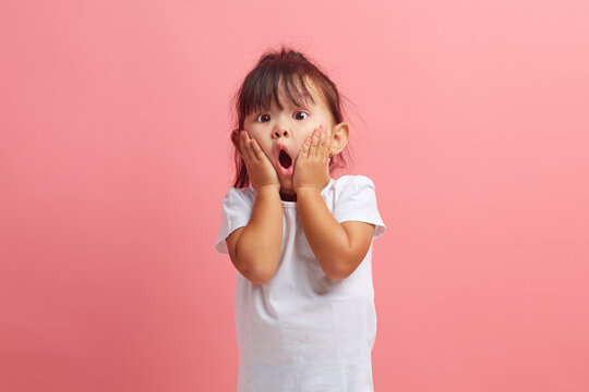 Amazed horrified little three years old girl keeps mouth opened, hands on cheeks, being shocked stares at camera, isolated over pink background. Kid expresses great surprisement.