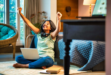 Happy teenage Indian asian student celebrating exam results at home with books, laptop on the bed