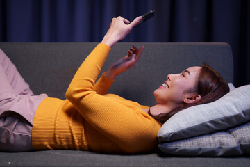 Young woman lying comfortably on a sofa, using her smartphone with a cheerful expression, enjoying a relaxing evening at home while browsing social media and connecting online