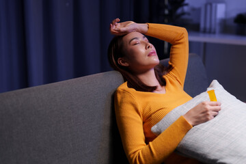 Young asian woman lying on sofa in living room at night, holding medicine, feeling unwell,...