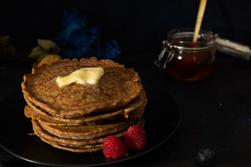 Stacked pancakes made with whole wheat flour with melted butter on top and raspberries on the side on a black plate against a dark background. Homemade food, Dark food
