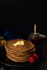 Vertical image of stacked pancakes made with whole wheat flour with melted butter on top and raspberries on the side on a black plate against a dark background. Homemade food, Dark food