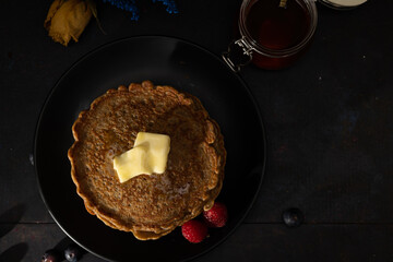 Top view of stacked pancakes made with whole wheat flour with melted butter on top and raspberries on the side on a black plate against a dark background. Homemade food, Dark food