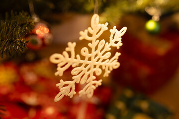 Close-up of a delicate white snowflake ornament hanging from a Christmas tree branch, with colorful decorations and wrapped gifts softly blurred in the background. 
