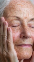 Fototapeta premium An elderly woman gently cradles her face with both hands, eyes closed, savoring a peaceful moment of reflection. Soft light highlights the warmth of her skin and tranquility