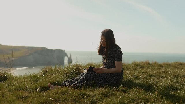On the ocean shore in Etretat. A girl reads the Bible in the open air, gratefully looking up to the sky. A woman holds the Bible in her hands and studies the word of God on the top of a cliff. Searchi