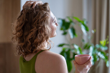 Serene female taking mindful pause with cup of tea, embracing slow living, relaxation. Happy woman feeling harmony, inner peace while reflecting on life gentle touching hair surrounded by houseplants