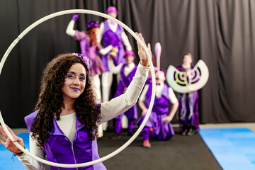 Young woman in purple costume holding white hula hoop with blurred circus performers in background, showcasing teamwork and entertainment