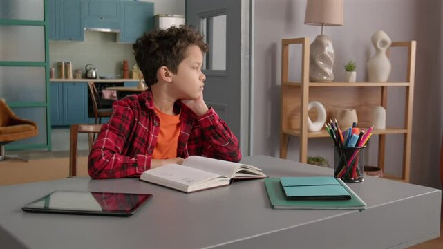 Cute school age black boy doing homework , reading textbook and having problems with understanding educational materials, expressing boredom and disinterest while learning at home.