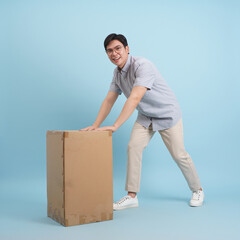 Young man pushing a cardboard box in a studio with a light blue background during daylight hours