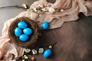 Bird's nest with blue Easter eggs, accented by white cherry blossom branches and soft beige fabric on brown textured background. Happy Easter greeting card template.