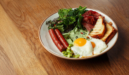 breakfast plate with fried eggs, grilled sausages, crispy bacon, toasted bread, fresh green salad. Healthy morning meal on a wooden table. copy space