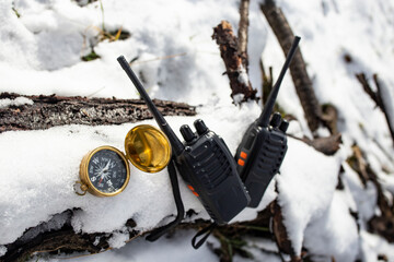 Two black walkie talkies on a tree log covered in frozen white snow next to a golden compass,...
