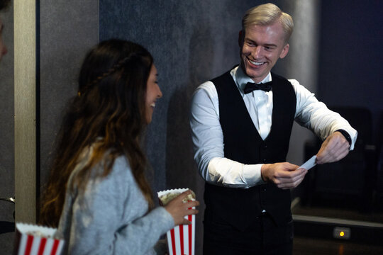 Smiling usher checking tickets at movie theater entrance with young woman holding popcorn